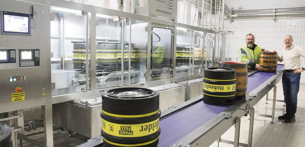 Die neue Fass-Abfüllanlage von Hohenfelder, hier präsentiert von Braumeister Heiko Krome (r.) und Mitarbeiter Thorsten Nobbe, kann von einem Mitarbeiter alleine bedient werden. Tatsächlich werden Fässer übrigens kopfüber gespült, damit u.a. Reinigungsmittel und Spülwasser auch durch Steigleitung und Ventil des Fasses zirkulieren können. Befüllt werden sie dann unter Druck: Die Flüssigkeit fließt sanft durch das Rohr zum Fassboden, während oben CO2 für Gegendruck sorgt. Foto: Schwade