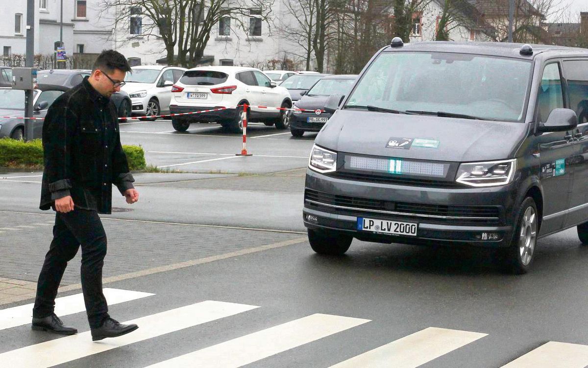 „Du kannst gehen, ich warte“: Mit einem eingeblendeten Pause-Zeichen an der Fahrzeugfront signalisiert der scheinbar autonom fahrende Hella-Bulli, dass seine Kameras den Fußgänger erkannt haben. Da kann Verkehrspsychologie-Student Jordan Raß ganz gelassen die Straße vor dem Lichtkanal überqueren. Fotos: Schwade