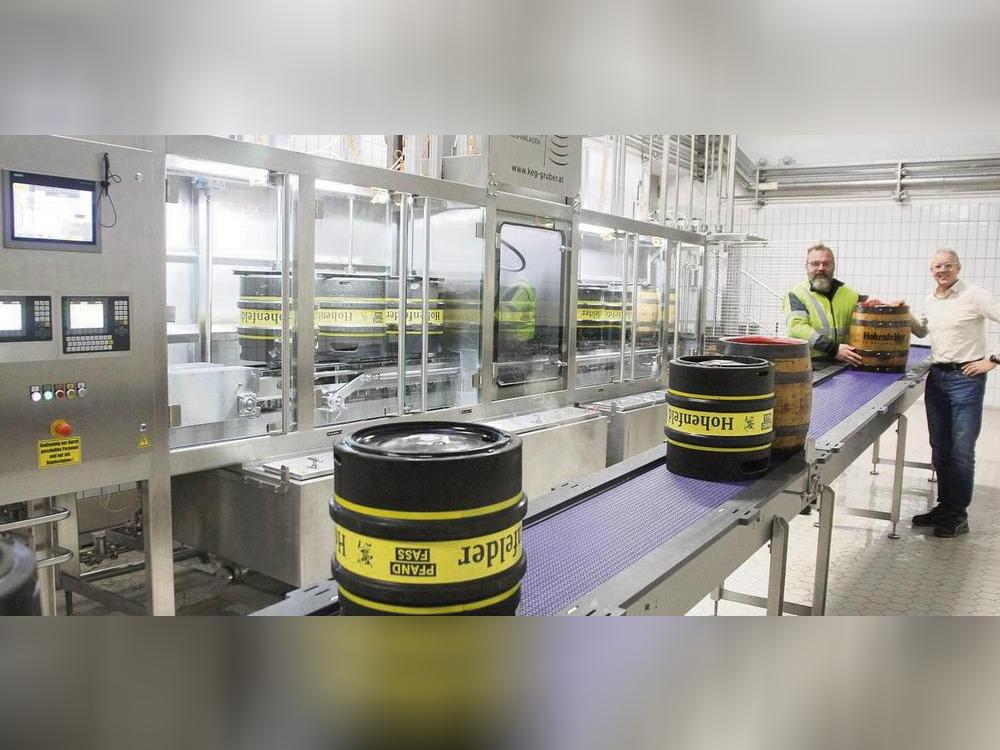 Die neue Fass-Abfüllanlage von Hohenfelder, hier präsentiert von Braumeister Heiko Krome (r.) und Mitarbeiter Thorsten Nobbe, kann von einem Mitarbeiter alleine bedient werden. Tatsächlich werden Fässer übrigens kopfüber gespült, damit u.a. Reinigungsmittel und Spülwasser auch durch Steigleitung und Ventil des Fasses zirkulieren können. Befüllt werden sie dann unter Druck: Die Flüssigkeit fließt sanft durch das Rohr zum Fassboden, während oben CO2 für Gegendruck sorgt. Foto: Schwade