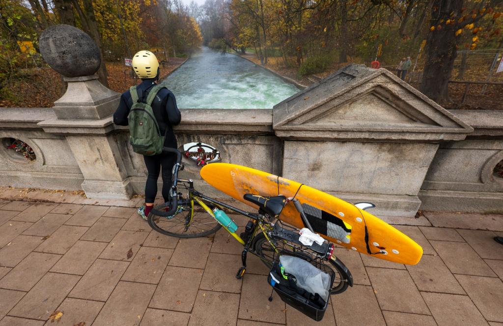 Ein Mann mit einem Surfboard an seinem Fahrrad schaut von einer Brücke auf die - nicht mehr vorhandene - Eisbachwelle im Englischen Garten. - Foto: Peter Kneffel/dpa